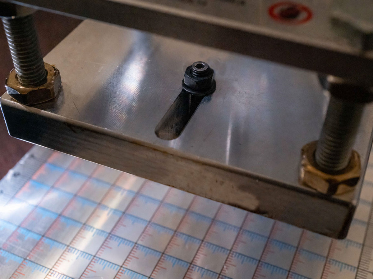 Close-up of a metal hot stamping press with threaded bolts and a sliding metal plate, positioned over a printed grid template.